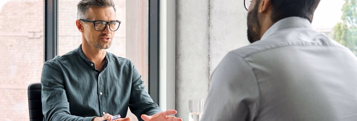 A man in glasses conversing with another man in an office setting.