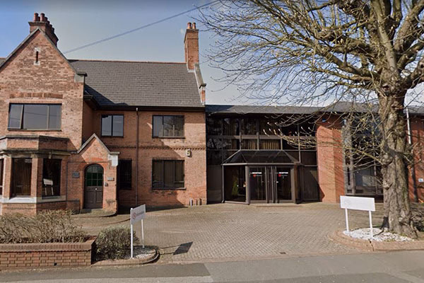 Market Location office, A two-story brick building with a sloped roof and multiple chimneys is adjacent to a glass entrance area. A tree and two white signboards are in the front of the building.